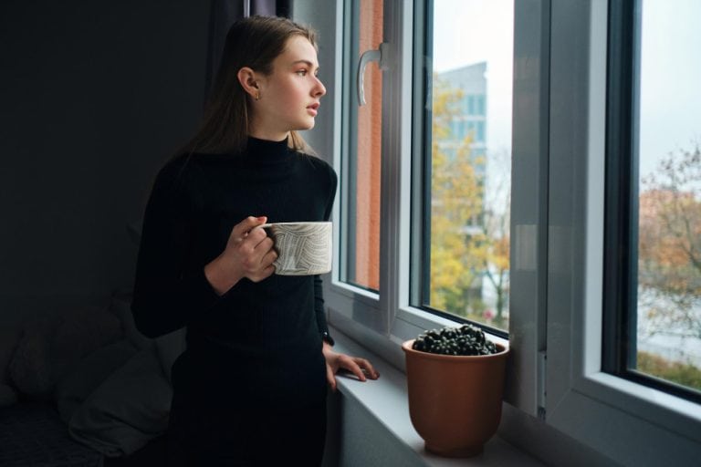 A young woman in a black turtleneck stands by a window, holding a ceramic mug and gazing thoughtfully outside. Natural light illuminates her face as autumn trees and a modern building appear in the background, suggesting a quiet moment of reflection or contemplation.