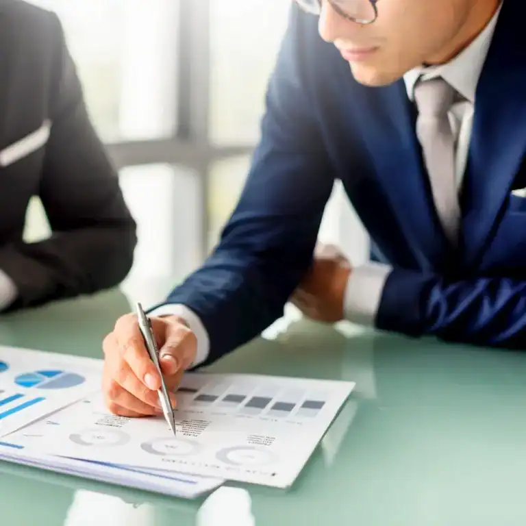 A businessman in a navy blue suit and glasses leans over a glass table, pointing at printed charts and graphs with a pen. The documents show various data visualizations, such as pie charts and bar graphs, indicating a professional discussion focused on financial performance or strategic planning.