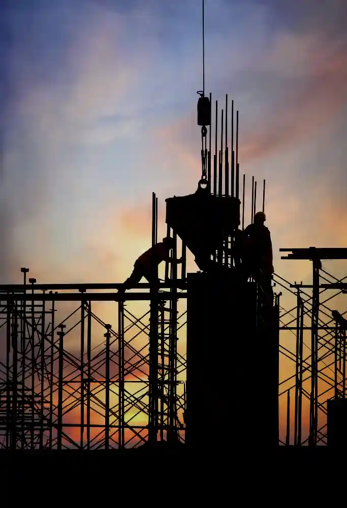 Silhouetted construction workers operate on scaffolding with a crane lifting a concrete bucket at sunset. The vibrant sky glows with orange, purple, and blue hues, highlighting the hard work and industrial setting—symbolic of labor-intensive industries like oil & gas or infrastructure.