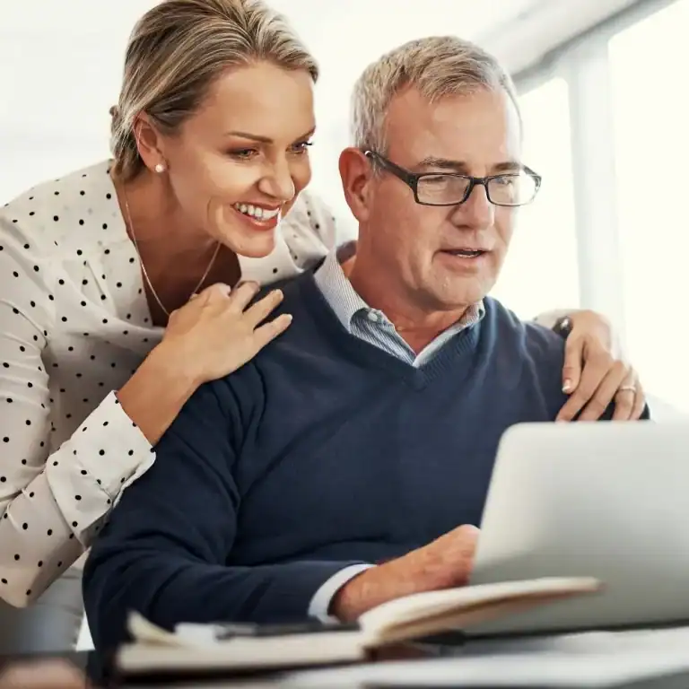 A smiling woman leans affectionately over the shoulder of a man wearing glasses and a blue sweater as he looks at a laptop screen. Both appear engaged and content, suggesting they are reviewing financial plans, retirement options, or personal investments together in a bright, relaxed home setting.