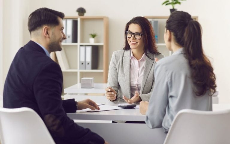 A professional woman in a light gray blazer and glasses smiles while talking to a man and a woman seated across the table in a modern office. She holds a pen and appears engaged in conversation, suggesting a financial planning or business advisory session. The setting is clean and welcoming, with shelves, folders, and plants in the background.