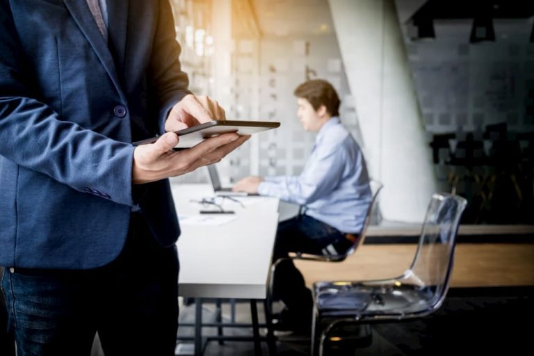 Businessman in a navy blazer using a digital tablet in a contemporary office environment, with a colleague working at a laptop in the background, highlighting modern workplace technology and collaborative settings.
