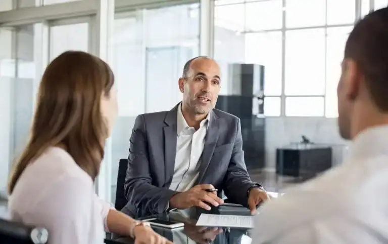 A financial advisor or business consultant in a gray suit sits across a glass table from a couple, explaining details from a document. The setting is a bright, modern office space with large windows, creating a professional yet approachable atmosphere for financial planning or investment discussion.