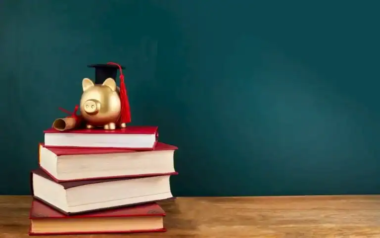 A golden piggy bank wearing a graduation cap sits atop a stack of thick red hardcover books, next to a tied diploma scroll. Set against a dark green chalkboard background on a wooden surface, the image symbolizes education savings, student loans, or financial planning for college