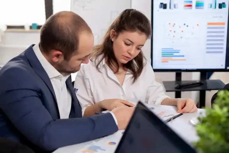 A man in a suit and a woman in a white shirt closely examine printed data charts during a meeting. Behind them, a large screen displays colorful graphs and analytics. The setting suggests a professional office environment focused on data analysis, business performance, or strategic planning.