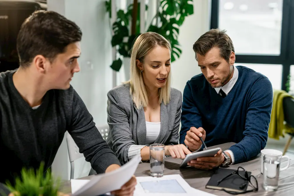 Three professionals sit at a modern office table engaged in a collaborative discussion. A woman in a gray blazer is pointing at a tablet held by a man in a blue sweater and tie, while another man looks on holding a document. Papers, water glasses, a notebook, and eyeglasses are spread across the table, emphasizing teamwork and strategic planning in a bright, plant-filled workspace.