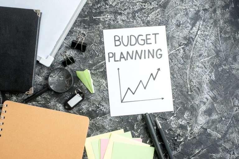 Flat lay of office supplies with a handwritten note titled ‘Budget Planning’ and a rising line graph on a textured desk surface
