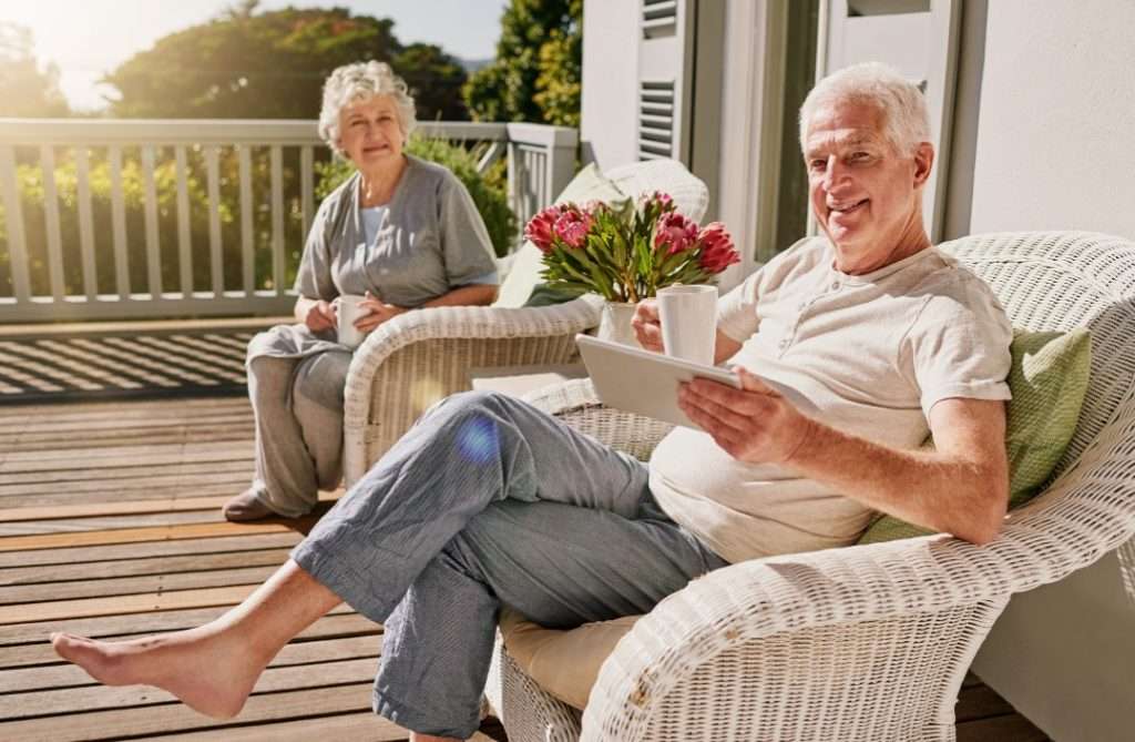 Smiling senior couple relaxing on a sunny patio with coffee and digital tablet, enjoying retirement and peaceful outdoor living