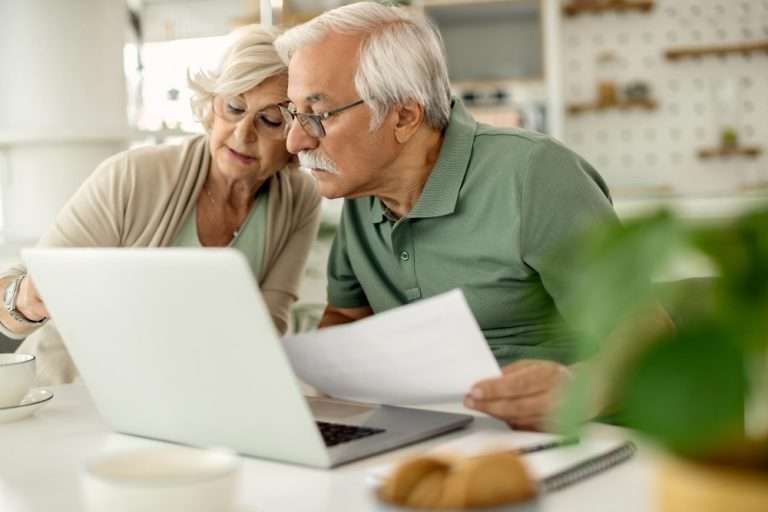 Senior couple reviewing financial documents together at home using a laptop, planning retirement or managing personal finances