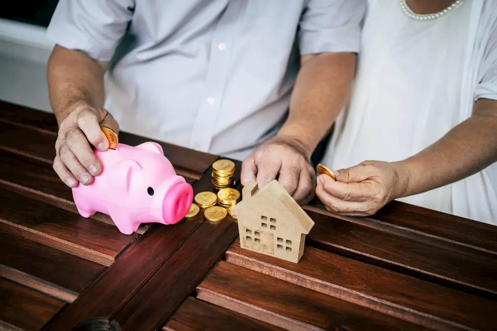 Senior couple saving money with gold coins, a pink piggy bank, and a wooden house model on a wooden table