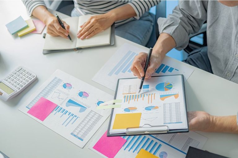 Two individuals examining financial graphs and data charts on papers spread out on a desk during a strategy meeting.